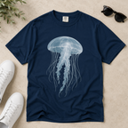 A navy blue short-sleeve t-shirt with a large white and light blue jellyfish graphic on the front, laid flat on a beige textured surface with white sneakers, sunglasses, and green leaves as props.