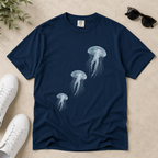 A navy blue short-sleeve t-shirt with a graphic print of three white jellyfish, styled in a flat-lay arrangement with white sneakers, sunglasses, and green leaves on a textured beige background.