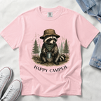 A light pink short-sleeve t-shirt laid flat on a white fabric background, featuring a graphic of a raccoon wearing a brown hat and green bandana while holding a mug, with the text 'HAPPY CAMPER' below. White sneakers and a denim jacket are partially visible as styling props.