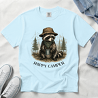 A light blue short-sleeve t-shirt laid flat on a white fabric background, featuring a graphic of a raccoon wearing a hat and scarf while holding a mug, with the text 'HAPPY CAMPER' below. White sneakers and a denim jacket are partially visible as styling props.