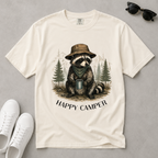 An off-white graphic t-shirt laid flat on a gray surface, styled with white sneakers and sunglasses. The t-shirt features a graphic of a raccoon wearing a hat and bandana, holding a mug in a forest, with the text 'HAPPY CAMPER' below.