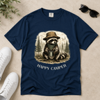 A navy blue short-sleeve t-shirt laid flat on a light beige textured surface, featuring a graphic of a raccoon wearing a hat and bandana while holding a mug in a forest, with the text 'HAPPY CAMPER' below. White sneakers, sunglasses, and a small plant sprig are styled around the shirt.