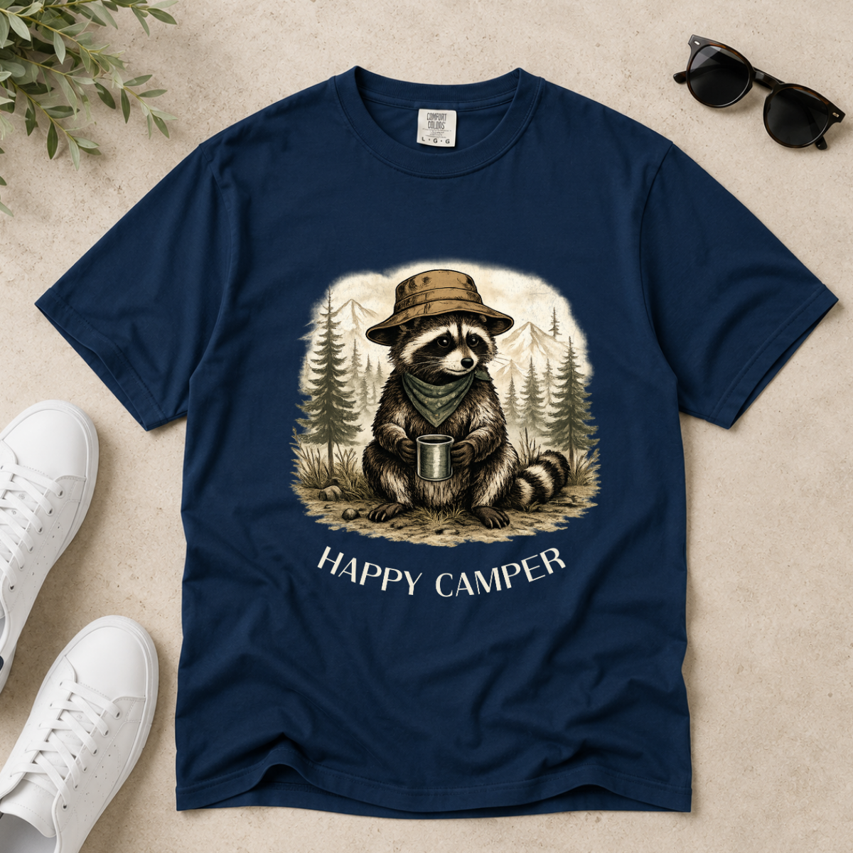 A navy blue short-sleeve t-shirt laid flat on a light beige textured surface, featuring a graphic of a raccoon wearing a hat and bandana while holding a mug in a forest, with the text 'HAPPY CAMPER' below. White sneakers, sunglasses, and a small plant sprig are styled around the shirt.
