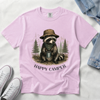 A light pink short-sleeve t-shirt laid flat on a white fabric background, featuring a graphic of a raccoon wearing a hat and holding a mug with the text 'HAPPY CAMPER' below. White sneakers and a denim jacket are partially visible as styling props.
