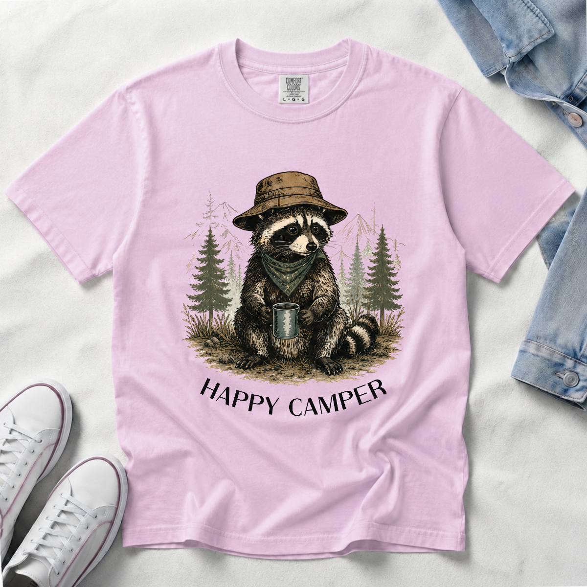 A light pink short-sleeve t-shirt laid flat on a white fabric background, featuring a graphic of a raccoon wearing a hat and holding a mug with the text 'HAPPY CAMPER' below. White sneakers and a denim jacket are partially visible as styling props.