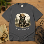A dark gray crewneck t-shirt featuring a graphic of a raccoon wearing a hat and scarf while holding a mug, with the text 'HAPPY CAMPER' below. The shirt is displayed in a flat-lay arrangement on a wooden surface alongside hiking boots, a compass, a map, and pine branches.