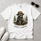 A white short-sleeve graphic T-shirt laid flat on a light gray surface, featuring a raccoon illustration wearing a hat and scarf while holding a mug, with the text 'HAPPY CAMPER' below. White sneakers, sunglasses
