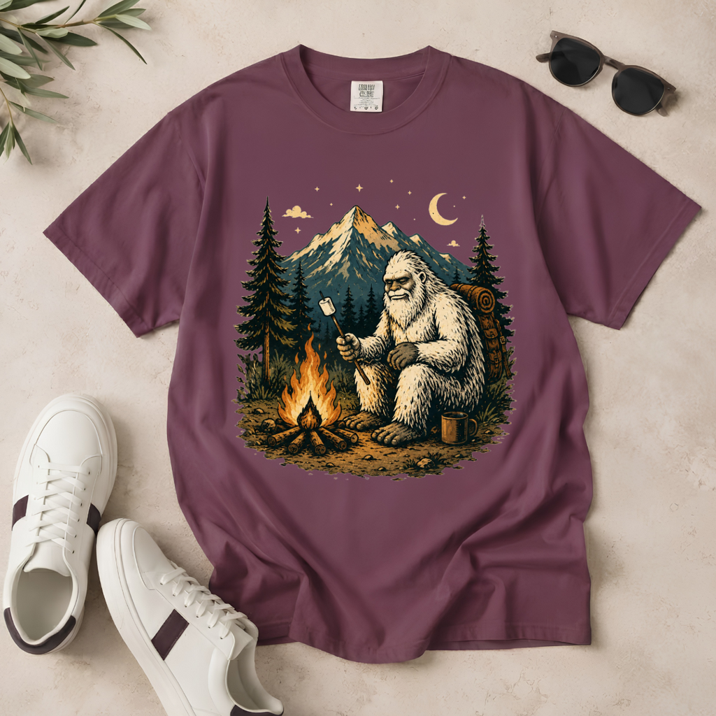 A mauve-purple short-sleeve t-shirt laid flat on a light beige surface, featuring a large graphic print of a Bigfoot figure roasting a marshmallow by a campfire at night, with mountains and pine trees in the background. White sneakers, black sunglasses, and a small plant sprig are styled around the shirt.