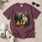 A mauve-purple short-sleeve t-shirt laid flat on a light beige surface, featuring a large graphic print of a Bigfoot figure roasting a marshmallow by a campfire at night, with mountains and pine trees in the background. White sneakers, black sunglasses, and a small plant sprig are styled around the shirt.
