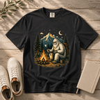 A black graphic t-shirt laid flat on a rustic wooden surface, featuring a detailed illustration of a Yeti sitting by a campfire under a mountain night sky. White sneakers and a notebook are styled around the shirt.