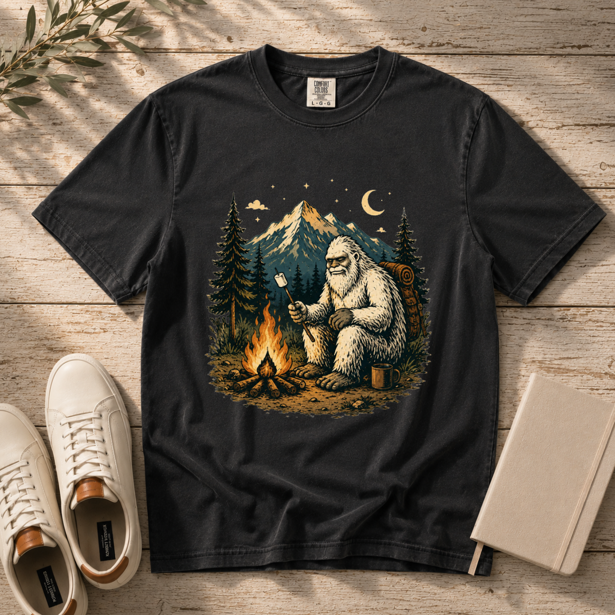 A black graphic t-shirt laid flat on a rustic wooden surface, featuring a detailed illustration of a Yeti sitting by a campfire under a mountain night sky. White sneakers and a notebook are styled around the shirt.