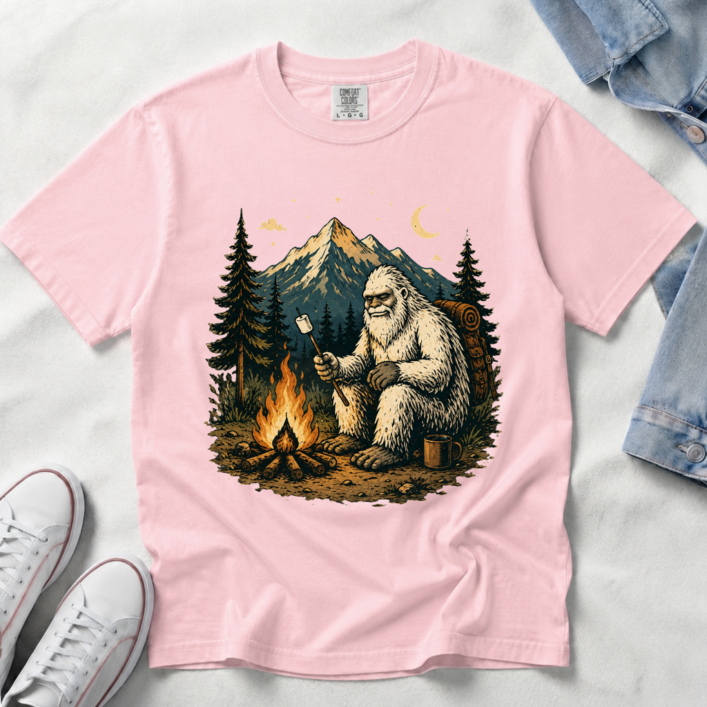 A blossom, light pink short-sleeve t-shirt laid flat on a white fabric background, featuring a large graphic print of a Bigfoot figure sitting by a campfire in front of mountains and pine trees. White sneakers and a folded pair of blue jeans are partially visible around the shirt as styling props.