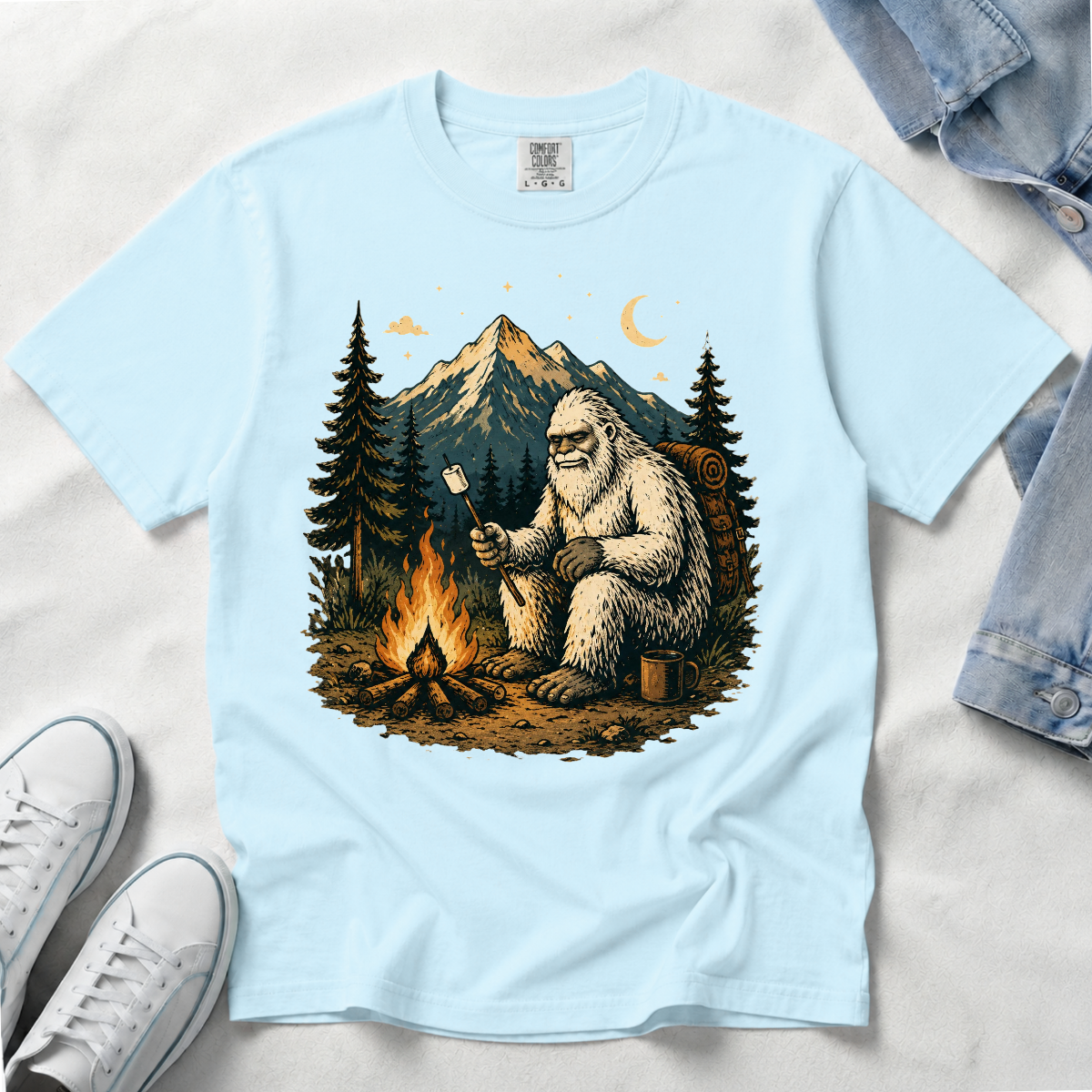 A light blue short-sleeve t-shirt laid flat on a white sand background, featuring a large graphic print of a Bigfoot figure sitting by a campfire with a marshmallow, surrounded by pine trees and a mountain under a crescent moon. White sneakers and a denim jacket are partially visible as styling props.