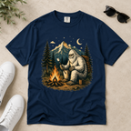 A navy blue short-sleeve t-shirt with a detailed graphic of a Yeti sitting by a campfire in front of a mountain. The shirt is displayed in a flat-lay arrangement on a textured beige surface, styled with white sneakers, sunglasses, and a sprig of green leaves.