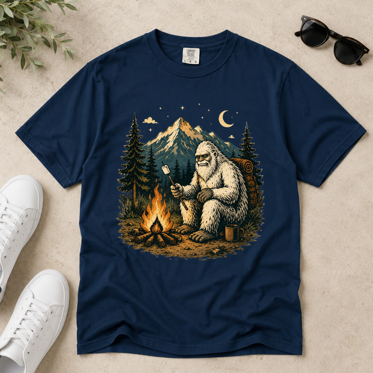 A navy blue short-sleeve t-shirt with a detailed graphic of a Yeti sitting by a campfire in front of a mountain. The shirt is displayed in a flat-lay arrangement on a textured beige surface, styled with white sneakers, sunglasses, and a sprig of green leaves.