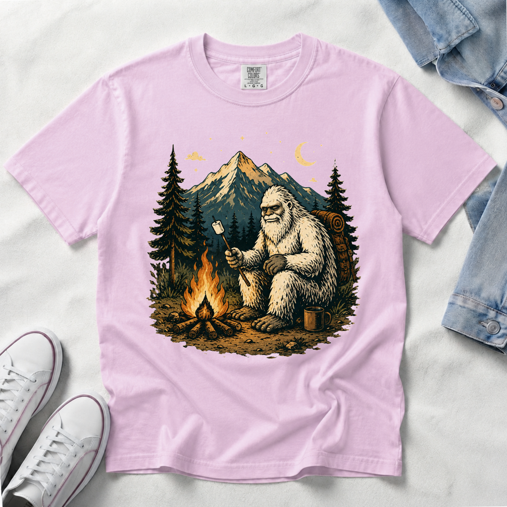 An orchid, light pink short-sleeve t-shirt laid flat on a white fabric background, featuring a large graphic print of a Bigfoot figure sitting by a campfire in front of mountains and pine trees. White sneakers and a folded denim jacket are partially visible as styling props.