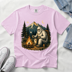 An orchid, light pink short-sleeve t-shirt laid flat on a white fabric background, featuring a large graphic print of a Bigfoot figure sitting by a campfire in front of mountains and pine trees. White sneakers and a folded denim jacket are partially visible as styling props.