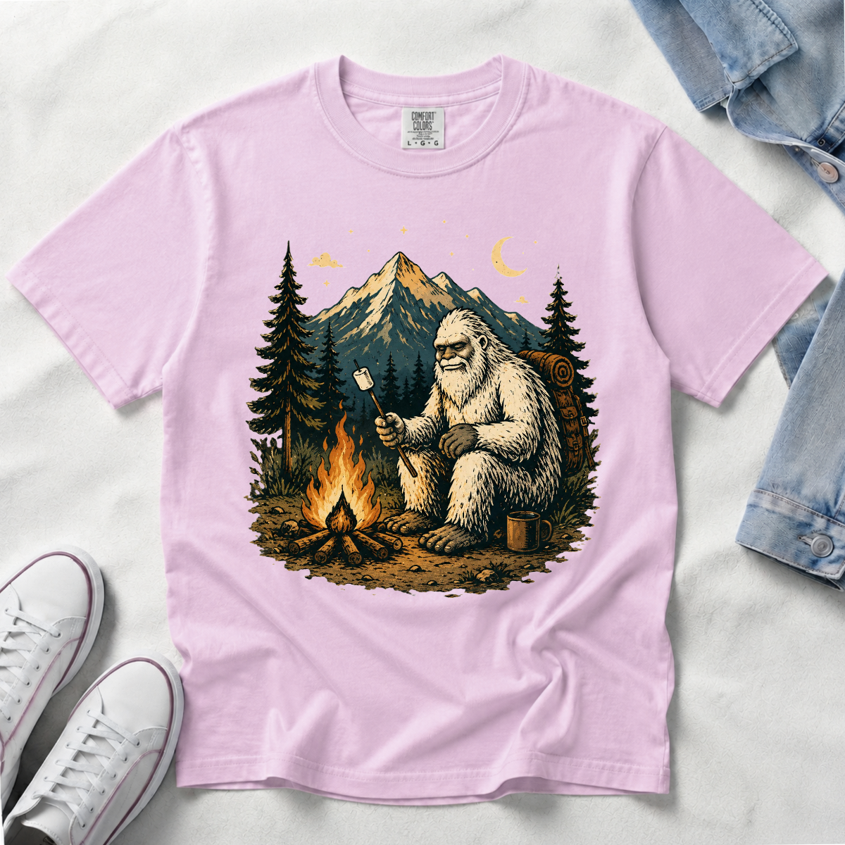 An orchid, light pink short-sleeve t-shirt laid flat on a white fabric background, featuring a large graphic print of a Bigfoot figure sitting by a campfire in front of mountains and pine trees. White sneakers and a folded denim jacket are partially visible as styling props.