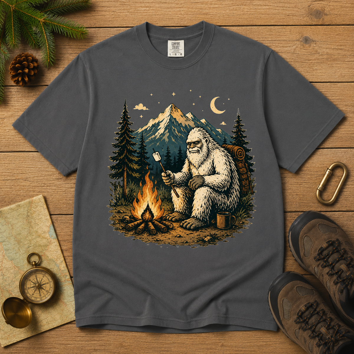 A dark gray crewneck t-shirt laid flat on a wooden surface, featuring a detailed graphic of a Yeti sitting by a campfire with a marshmallow. The shirt is styled with outdoor props including pine branches, a compass, a map, a brass key, and hiking boots.