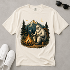 An off-white short-sleeve t-shirt with a detailed graphic of a Bigfoot sitting by a campfire in front of mountains and pine trees. The shirt is styled in a flat-lay arrangement on a gray background, surrounded by white sneakers, sunglasses, and a small moon graphic.