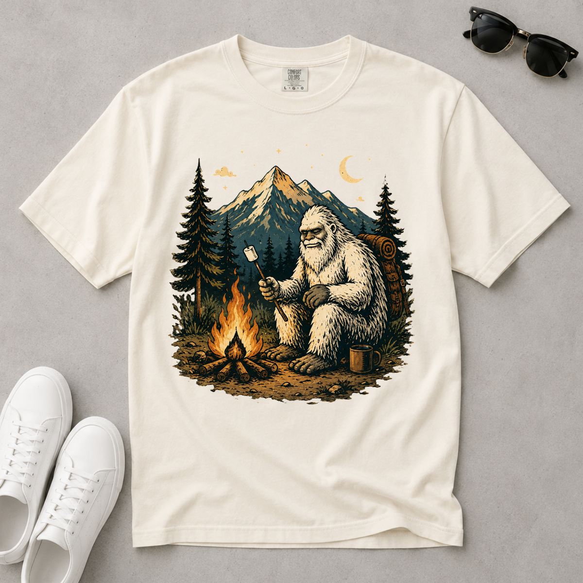 An off-white short-sleeve t-shirt with a detailed graphic of a Bigfoot sitting by a campfire in front of mountains and pine trees. The shirt is styled in a flat-lay arrangement on a gray background, surrounded by white sneakers, sunglasses, and a small moon graphic.