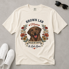 An off-white short-sleeve graphic t-shirt laid flat on a gray surface, featuring a brown Labrador retriever illustration surrounded by flowers and the text 'BROWN LAB Mom FUELED BY COFFEE & Lab Love'. White sneakers and black sunglasses are styled around the shirt.