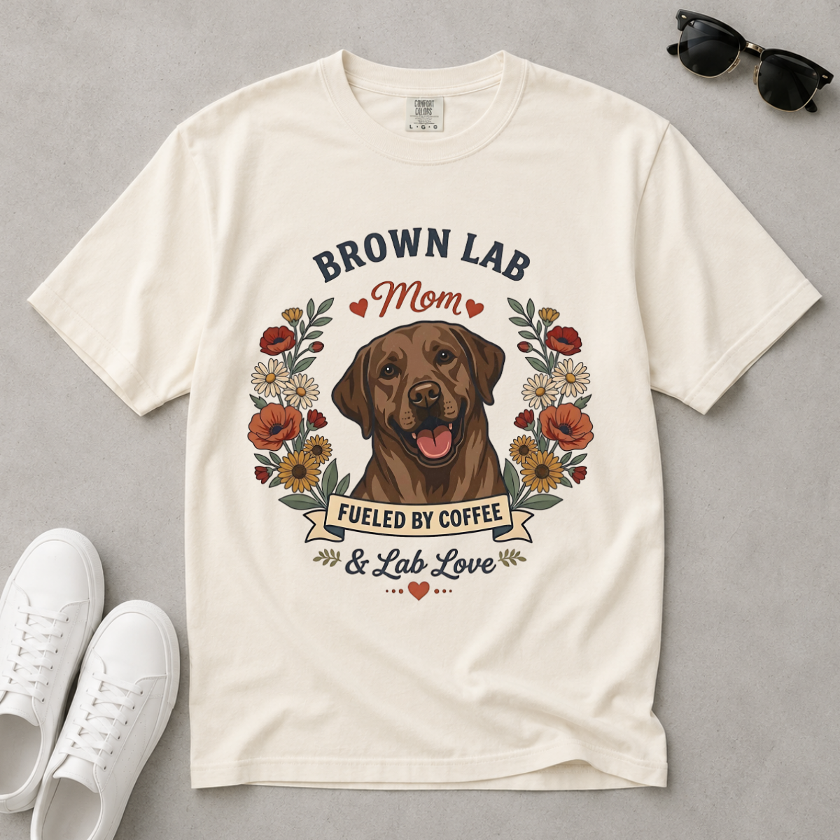 An off-white short-sleeve graphic t-shirt laid flat on a gray surface, featuring a brown Labrador retriever illustration surrounded by flowers and the text 'BROWN LAB Mom FUELED BY COFFEE & Lab Love'. White sneakers and black sunglasses are styled around the shirt.