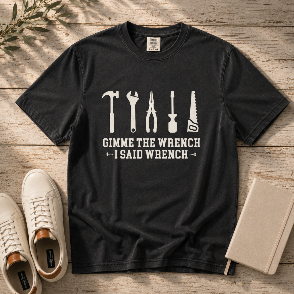 A black graphic t-shirt laid flat on a rustic wooden surface, styled with white sneakers, a notebook, and a sprig of greenery. The t-shirt features white line-art icons of various tools (hammer, wrench, pliers, screwdriver, saw) above the text 'GIMME THE WRENCH I SAID WRENCH'.