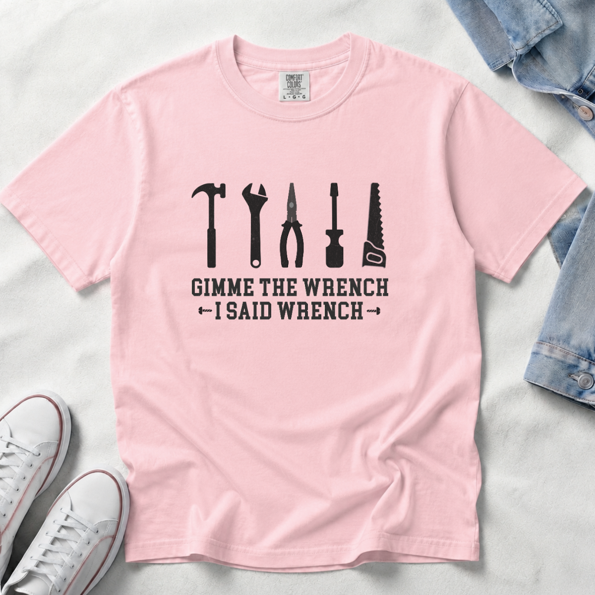 A light pink short-sleeve t-shirt with a black graphic of various tools and the text 'GIMME THE WRENCH - I SAID WRENCH', laid flat on a white fabric background with jeans and white sneakers as styling props.