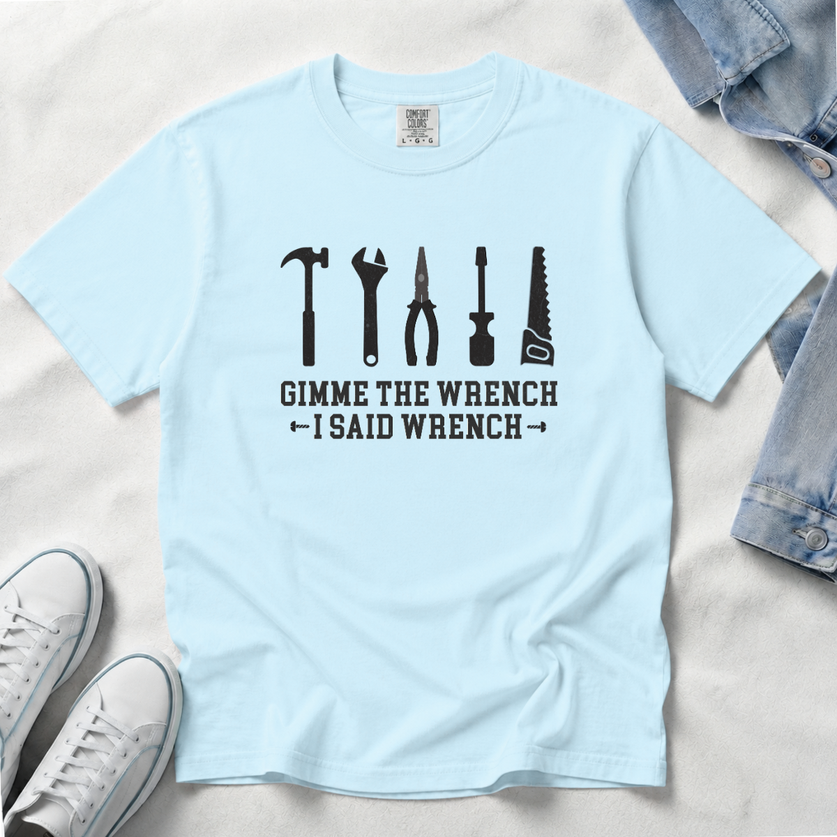 A light blue crewneck t-shirt with a black graphic of various tools and the text 'GIMME THE WRENCH I SAID WRENCH', laid flat on a white fabric background with jeans and sneakers as props.