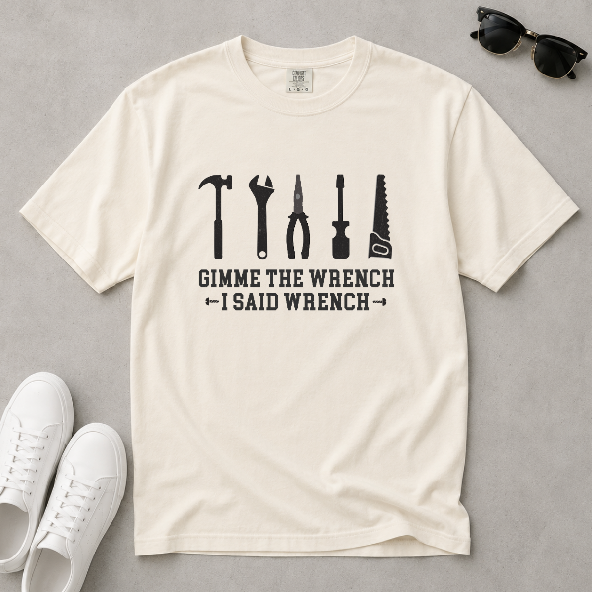 An off-white crewneck t-shirt with a black graphic of six hand tools and the text 'GIMME THE WRENCH I SAID WRENCH' laid flat on a gray background with white sneakers and sunglasses as props.
