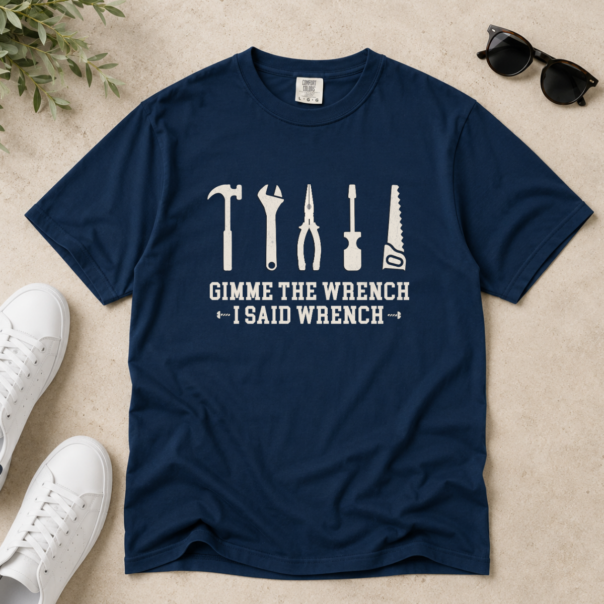 A navy blue short-sleeve t-shirt with a white graphic of various hand tools and the text 'GIMME THE WRENCH I SAID WRENCH', laid flat on a light beige surface with white sneakers, sunglasses, and green leaves as props.