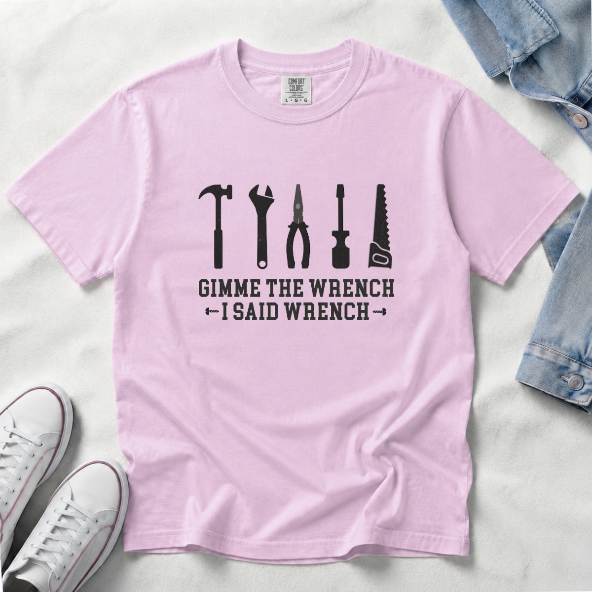 A light pink crewneck t-shirt with a black graphic of six tools (hammer, wrench, pliers, screwdriver, saw, nutcracker) and the text 'GIMME THE WRENCH I SAID WRENCH'. The shirt is laid flat on a white textured surface with styled props, including a denim jacket and white sneakers
