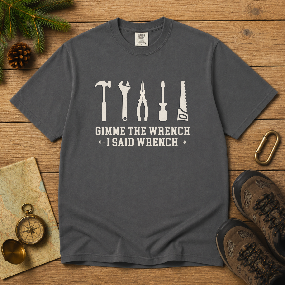 A dark gray crewneck t-shirt with a white graphic of six hand tools and the text 'GIMME THE WRENCH I SAID WRENCH', presented in a flat-lay arrangement on a wooden surface with hiking boots, a compass, and pine branches.