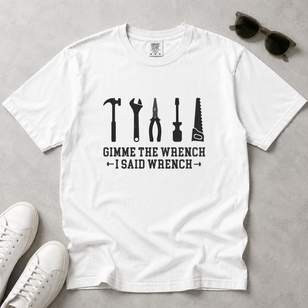 A white short-sleeve graphic T-shirt laid flat on a light gray surface, with black tool silhouettes and the text 'GIMME THE WRENCH I SAID WRENCH.' White sneakers and sunglasses are placed around it as styling props.