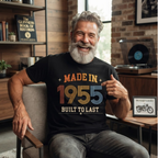A smiling older man with a gray beard sits in a chair wearing a black graphic T-shirt that reads 'MADE IN 1955 BUILT TO LAST' in orange, yellow, and blue lettering. The setting is a cozy indoor room with brick walls, shelves, and a record player in the background.