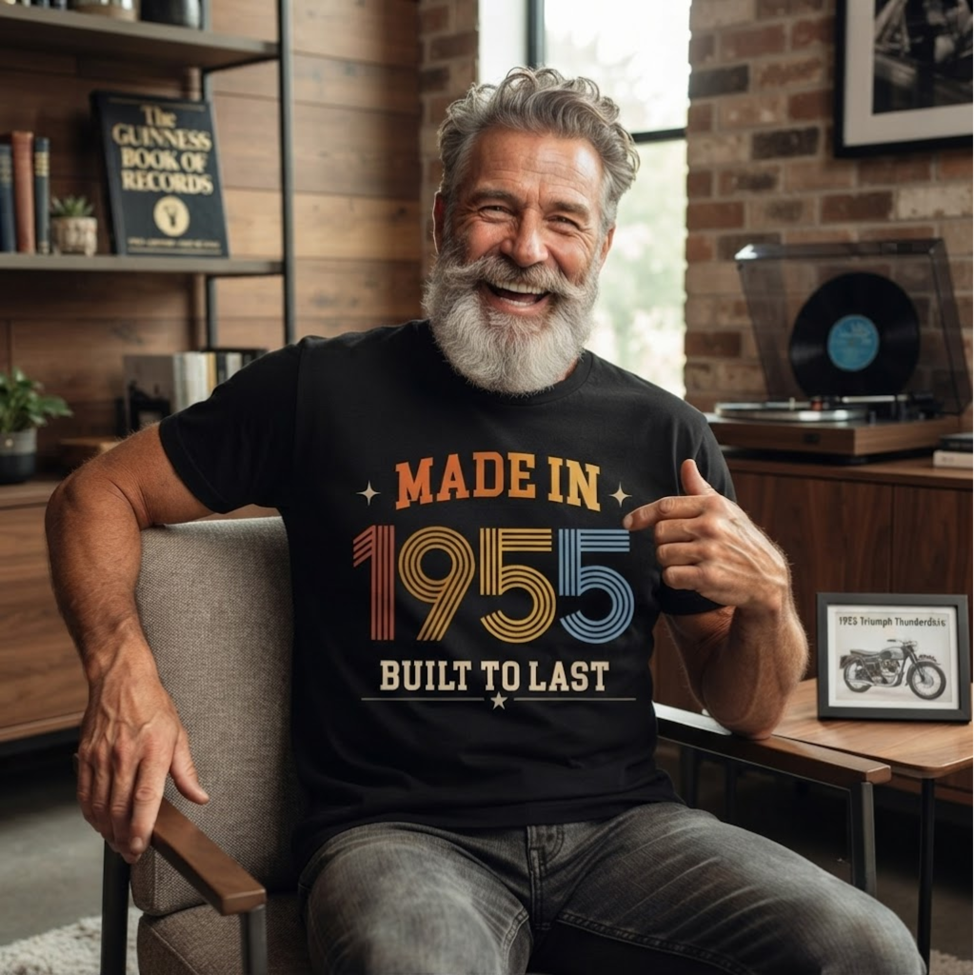 A smiling older man with a gray beard sits in a chair wearing a black graphic T-shirt that reads 'MADE IN 1955 BUILT TO LAST' in orange, yellow, and blue lettering. The setting is a cozy indoor room with brick walls, shelves, and a record player in the background.