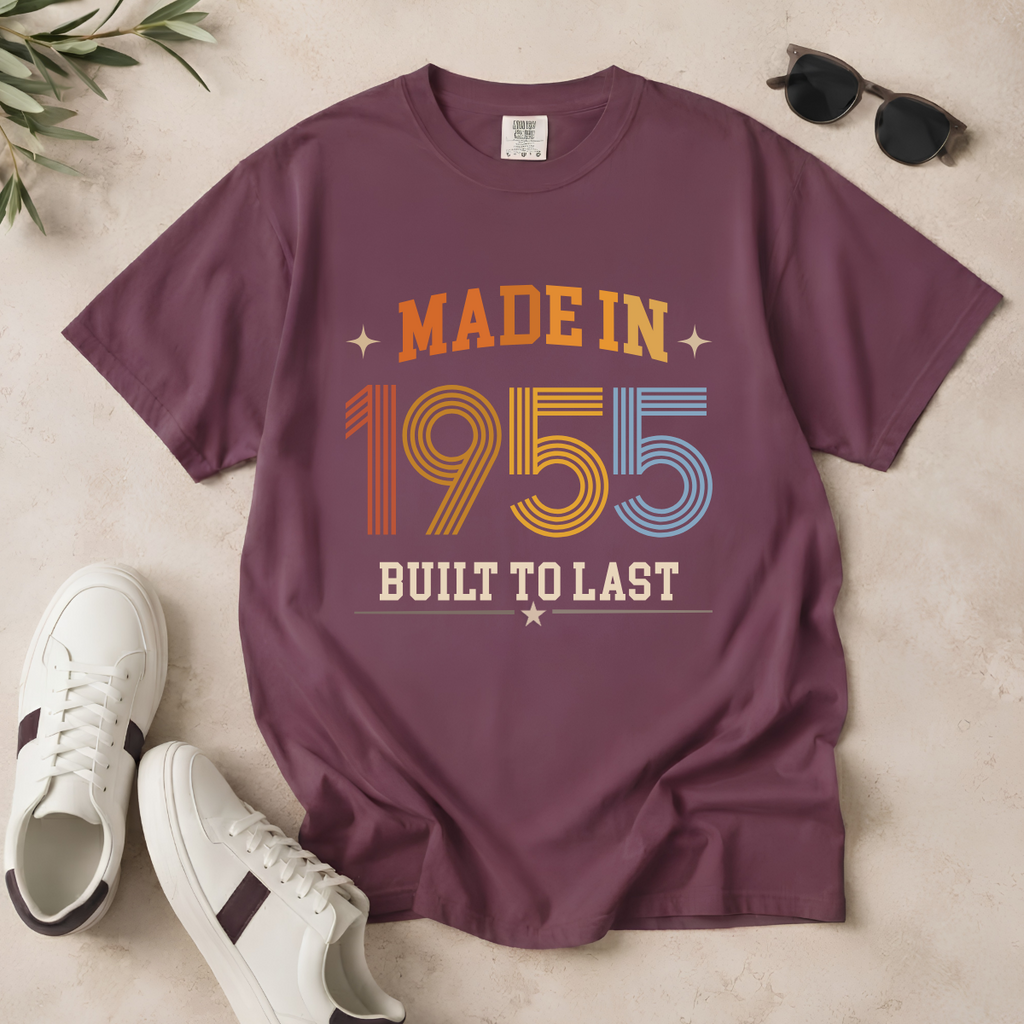 A mauve-purple short-sleeve graphic T-shirt laid flat on a light beige surface, styled with white sneakers, sunglasses, and a small plant sprig. The shirt reads 'MADE IN 1955 BUILT TO LAST' in retro orange, yellow, blue, and cream lettering. The perfect birthday gift for someone born in 1955.