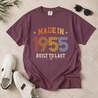 A mauve-purple short-sleeve graphic T-shirt laid flat on a light beige surface, styled with white sneakers, sunglasses, and a small plant sprig. The shirt reads 'MADE IN 1955 BUILT TO LAST' in retro orange, yellow, blue, and cream lettering. The perfect birthday gift for someone born in 1955.