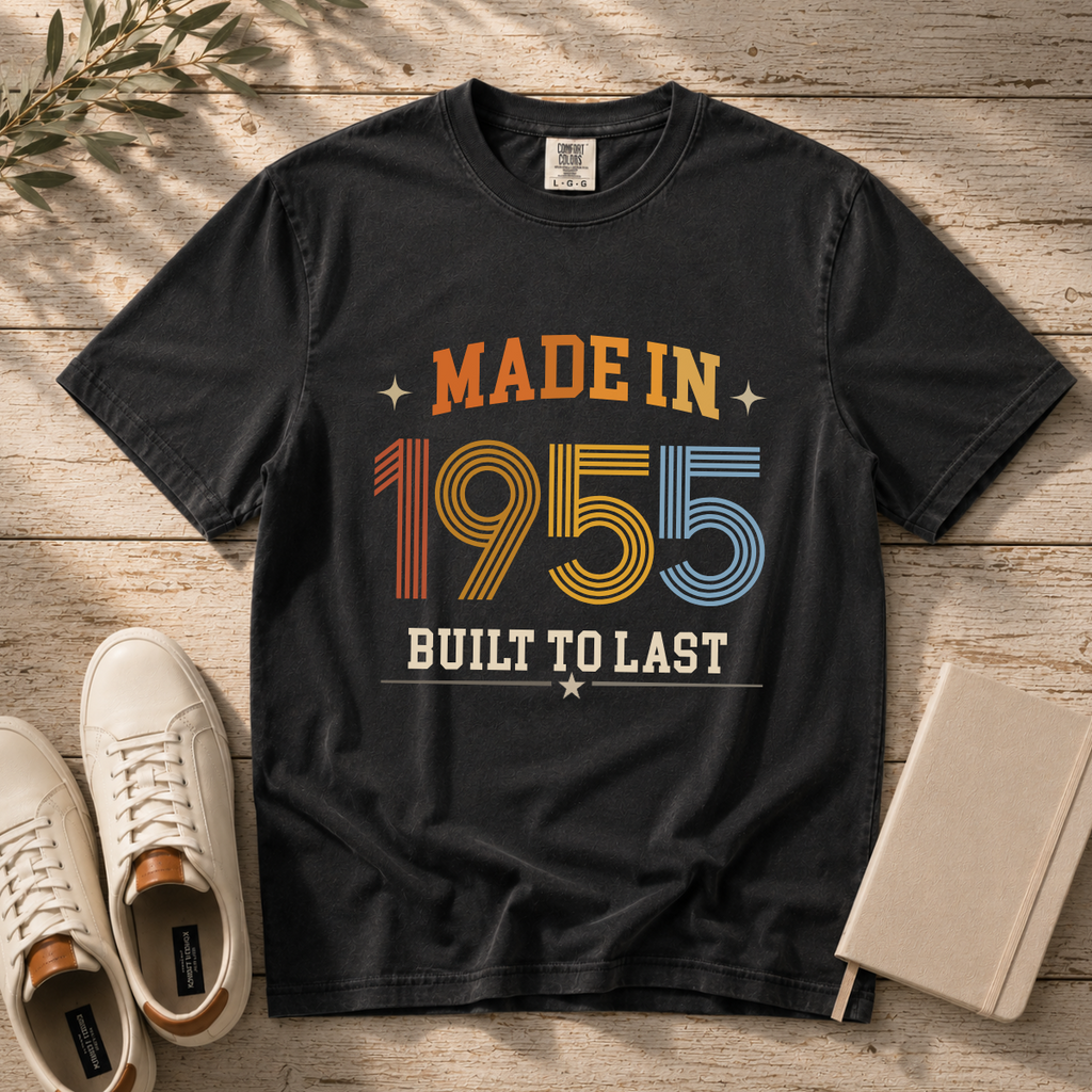 A black graphic t-shirt laid flat on a rustic wooden surface, styled with white sneakers, a notebook, and a sprig of greenery. The t-shirt features a retro-style graphic that reads 'MADE IN 1955 BUILT TO LAST' in orange, yellow, blue, and white colors. The perfect birthday gift for someone born in 1955.