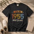 A black graphic t-shirt laid flat on a rustic wooden surface, styled with white sneakers, a notebook, and a sprig of greenery. The t-shirt features a retro-style graphic that reads 'MADE IN 1955 BUILT TO LAST' in orange, yellow, blue, and white colors. The perfect birthday gift for someone born in 1955.