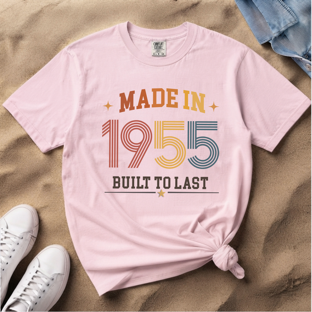 A light pink, blossom graphic T-shirt laid flat on sand, styled with white sneakers, blue jeans, and a knotted hem. The shirt reads 'MADE IN 1955 BUILT TO LAST' in retro multicolor lettering. The perfect birthday gift for someone born in 1955.