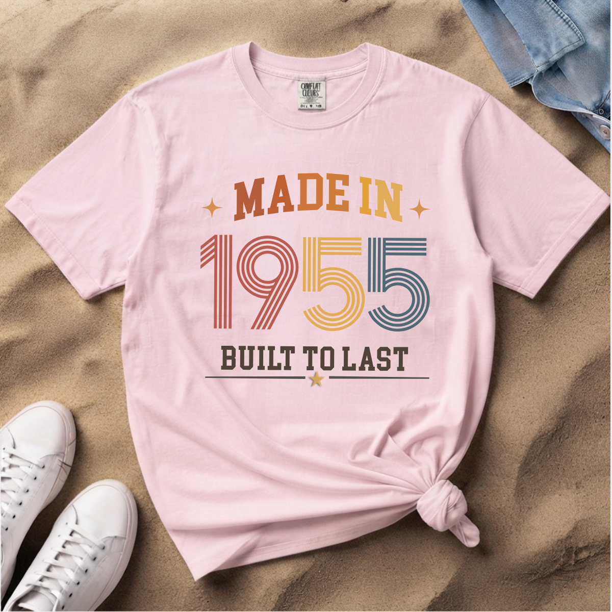 A light pink, blossom graphic T-shirt laid flat on sand, styled with white sneakers, blue jeans, and a knotted hem. The shirt reads 'MADE IN 1955 BUILT TO LAST' in retro multicolor lettering. The perfect birthday gift for someone born in 1955.