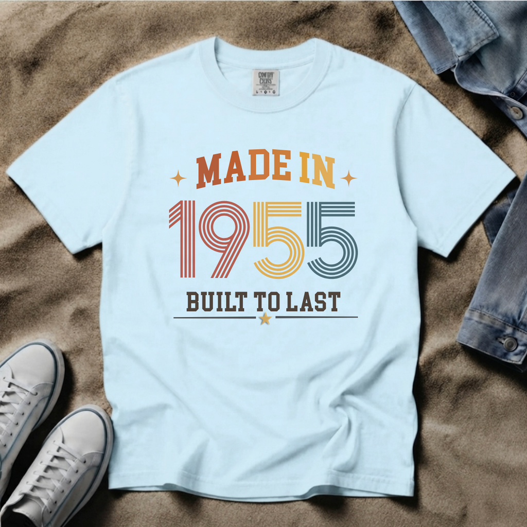 A light blue graphic t-shirt laid flat on a brown textured surface, styled with a pair of blue jeans and white sneakers. The t-shirt features a retro-style graphic that reads 'MADE IN 1955 BUILT TO LAST' in orange, yellow, and blue colors. The perfect birthday gift for someone born in 1955.