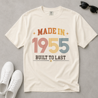 An off-white, ivory short-sleeve t-shirt with a retro-style graphic reading 'MADE IN 1955 BUILT TO LAST' in orange, red, yellow, and blue colors, styled in a flat-lay with white sneakers and sunglasses on a gray background. The perfect birthday gift for someone born in 1955.