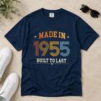 A navy blue graphic t-shirt laid flat on a light beige textured surface, styled with white sneakers, sunglasses, and green leaves. The t-shirt features a retro-style graphic that reads 'MADE IN 1955 BUILT TO LAST' in orange, yellow, and blue striped lettering. The perfect birthday gift for someone born in 1955.