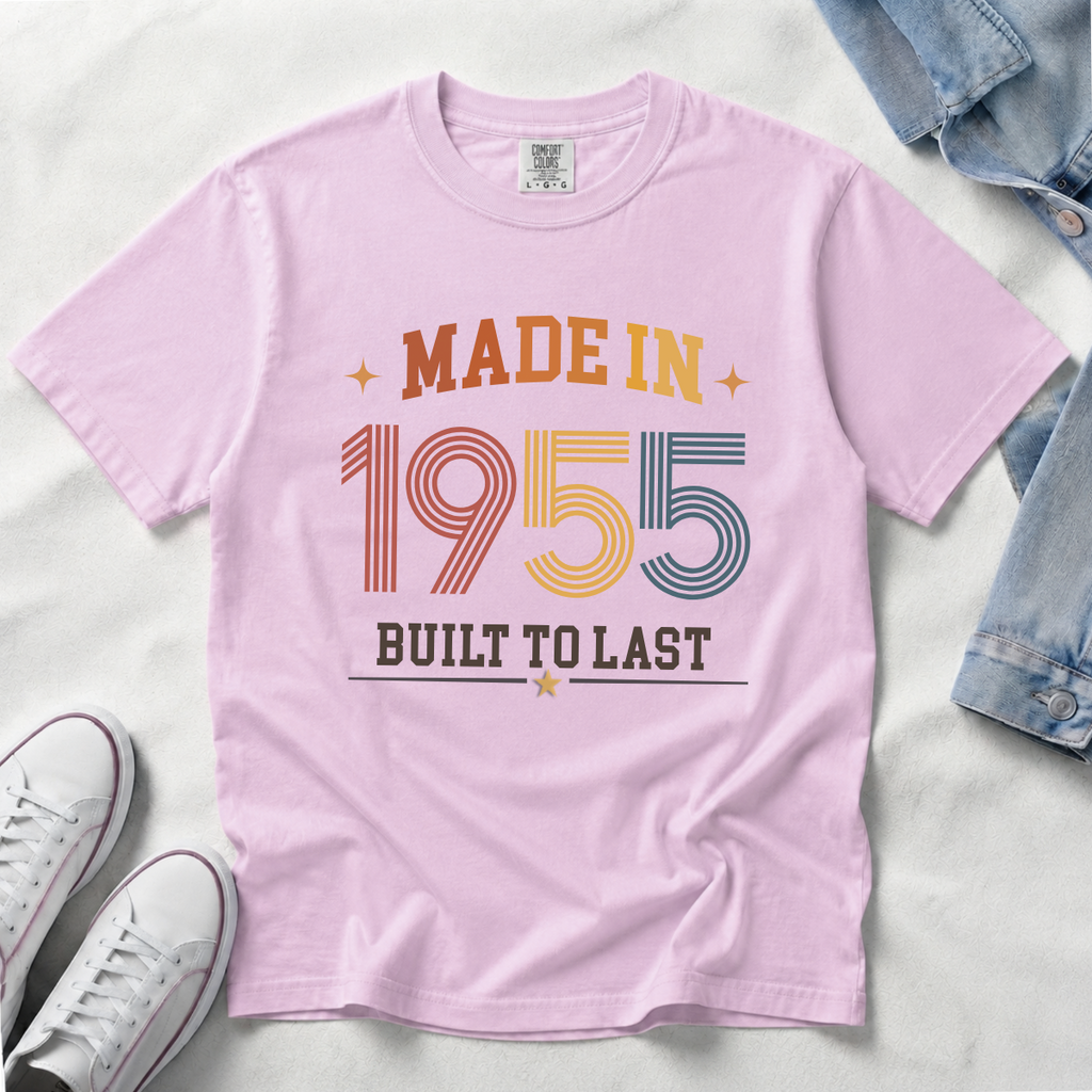 A light pink, orchid short-sleeve t-shirt laid flat on a white fabric background, featuring a retro-style graphic that reads 'MADE IN 1955 BUILT TO LAST' in orange, yellow, blue, and dark gray lettering. Styled flat-lay props include white sneakers at the bottom left, a denim jacket at the top right, and a white surface underneath. The perfect birthday gift for someone born in 1955.
