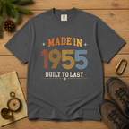 A dark gray , pepper graphic t-shirt laid flat on a wooden surface, surrounded by outdoor-themed props including pine branches, a pinecone, a compass, a map, a carabiner, and hiking boots. The t-shirt features a retro-style graphic that reads 'MADE IN 1955 BUILT TO LAST' in orange, yellow, blue, and white colors. The perfect birthday gift for someone born in 1955.