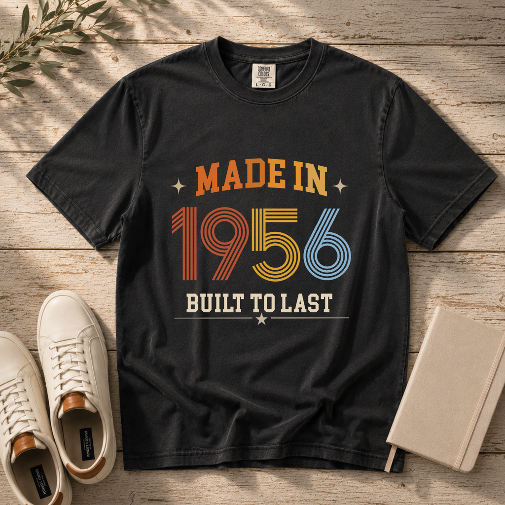 A black graphic t-shirt laid flat on a rustic wooden surface, styled with white sneakers, a notebook, and a sprig of greenery. The t-shirt features a retro-style graphic reading 'MADE IN 1956 BUILT TO LAST' in orange, yellow, and blue colors.