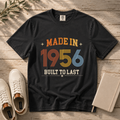 A black graphic t-shirt laid flat on a rustic wooden surface, styled with white sneakers, a notebook, and a sprig of greenery. The t-shirt features a retro-style graphic reading 'MADE IN 1956 BUILT TO LAST' in orange, yellow, and blue colors.